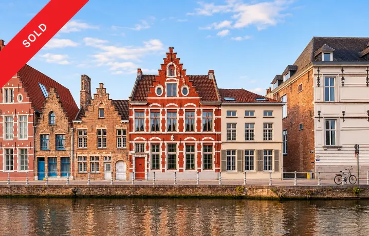Row of historic brick buildings along a canal in Belgium with a red "SOLD" banner in the corner under a blue sky.