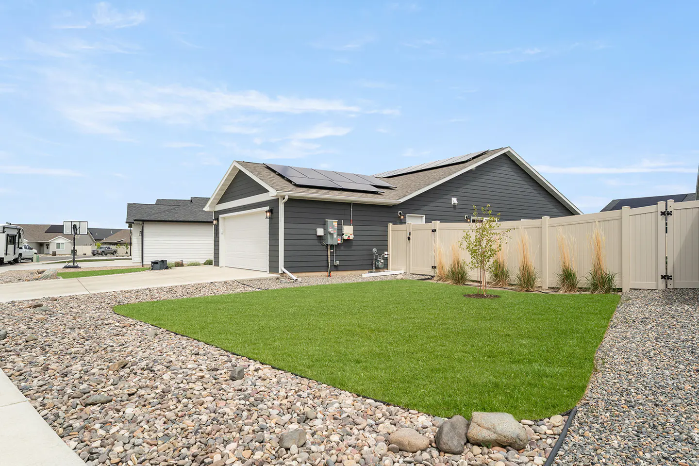 Exterior of a gray house with solar panels, a white garage door, and a fenced-in yard with green grass and rocks.