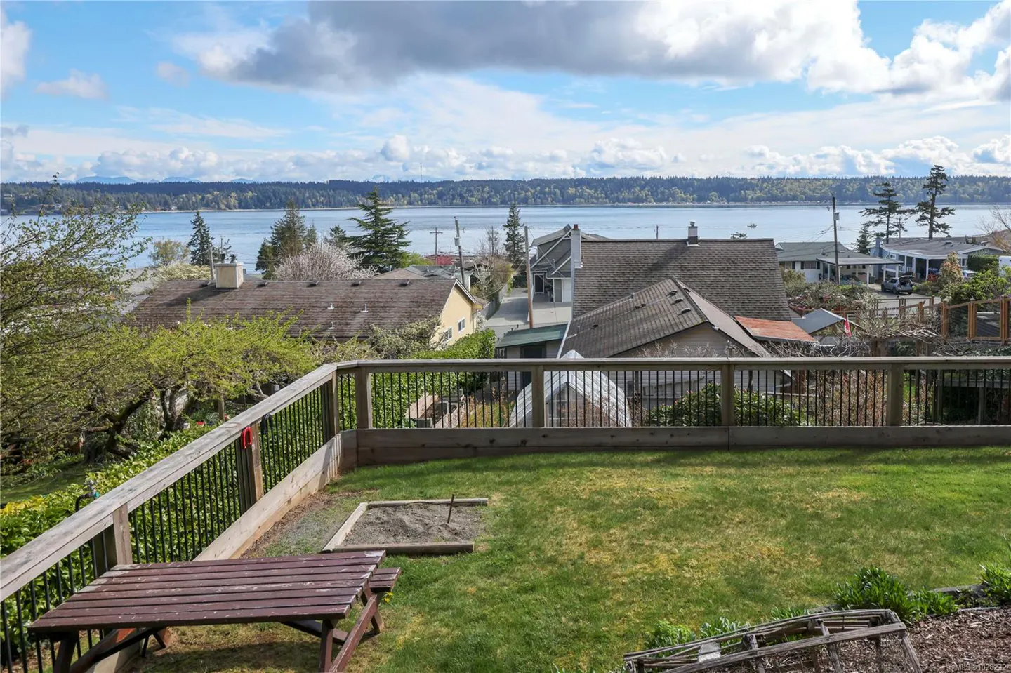 View from a deck with a picnic table and green lawn, overlooking houses, water, and a tree-lined shore under a cloudy sky.