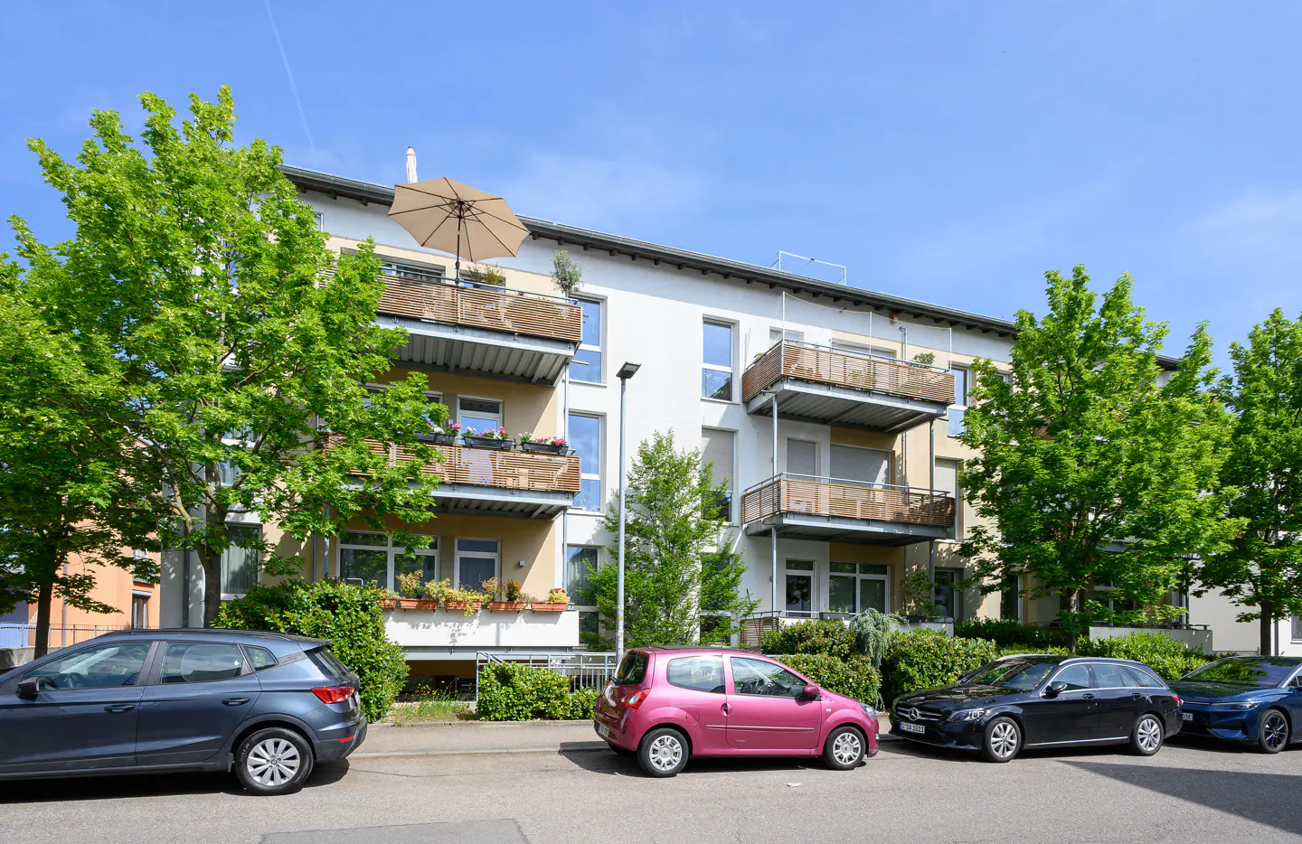 Apartment building with balconies, green trees, and parked cars on a sunny day.