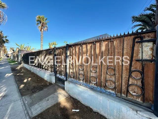 Exterior view of a property with a tall, ornate wooden and iron fence under a clear blue sky. Palm trees are visible in the background.