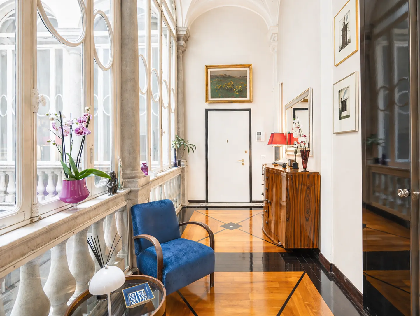 Hallway with wood floors, white walls, and large windows. A blue chair sits near a glass table with a white lamp. A wood dresser is against the wall.