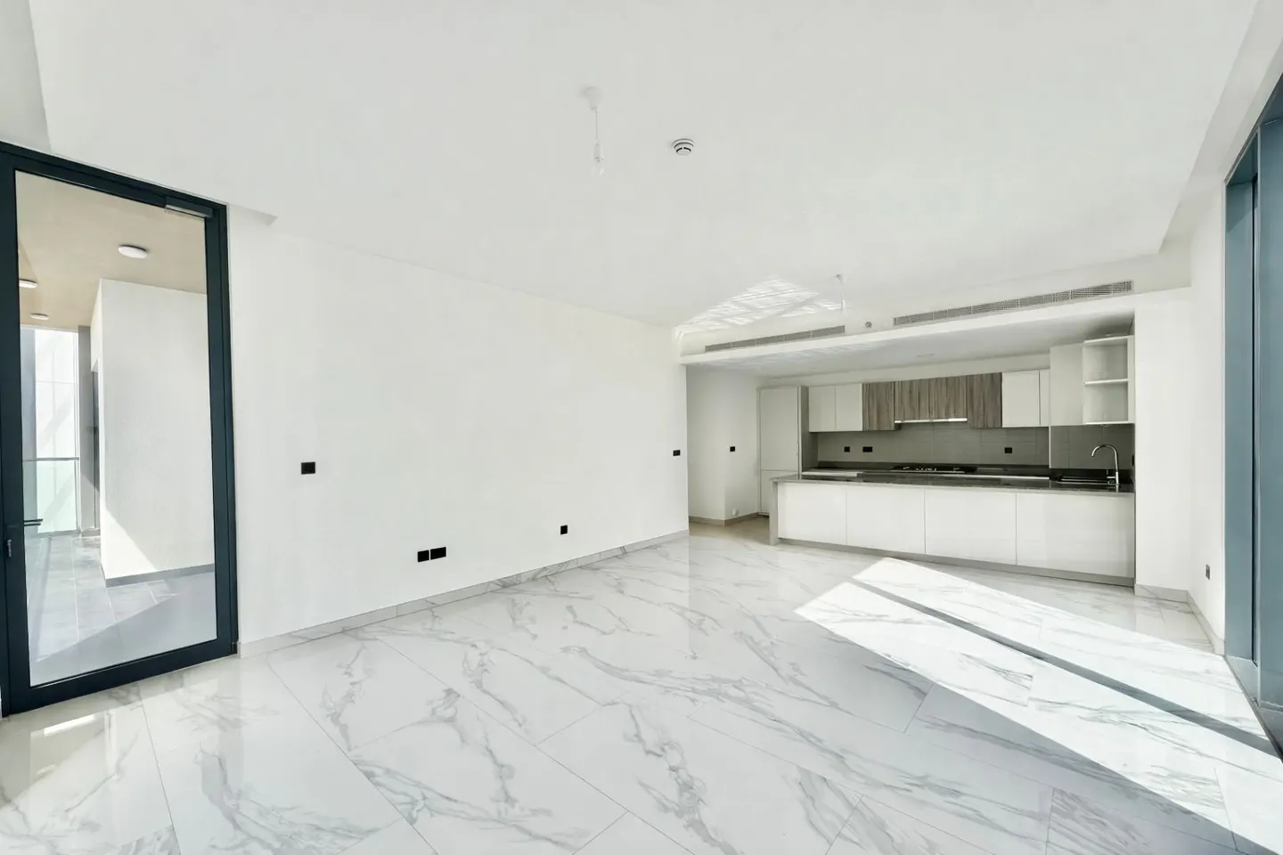 Bright, empty room with white walls, marble floors, and a modern kitchen in the background. A black-framed doorway is on the left.