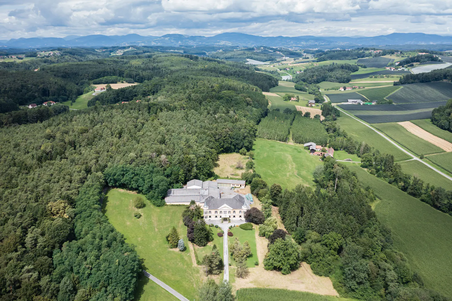 Aerial view of a large, light-yellow mansion surrounded by green lawns, trees, and forests, with mountains in the background under a cloudy sky.