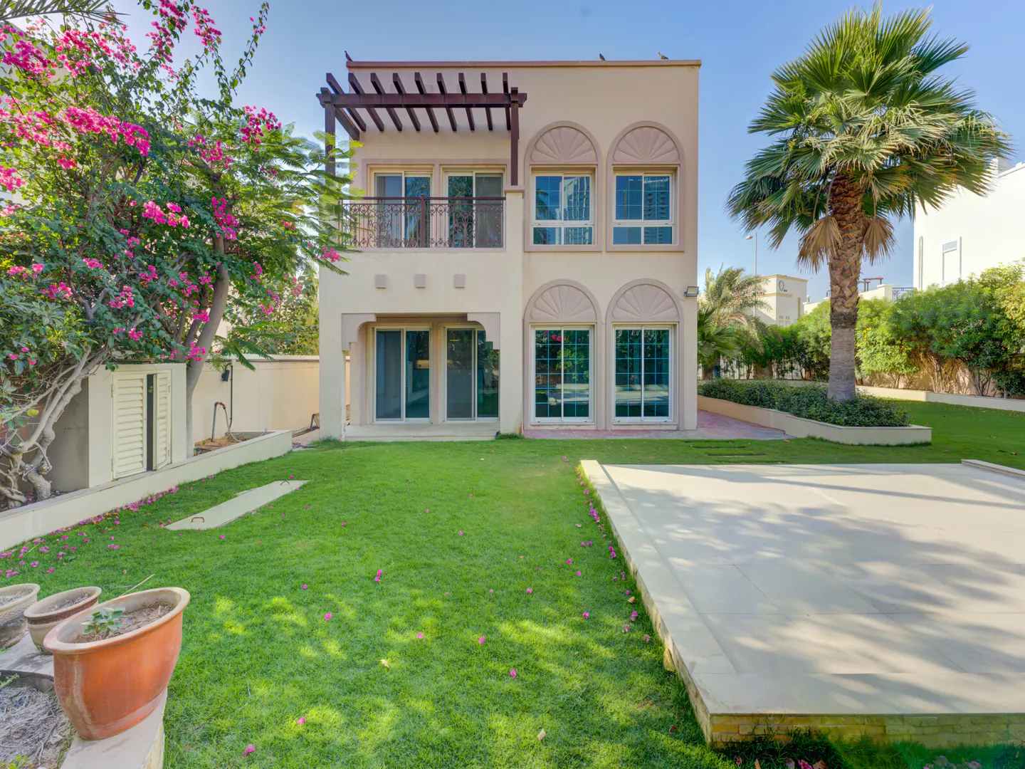 Two-story beige house with a green lawn, palm tree, and pink bougainvillea. The house has arched windows and a small balcony.