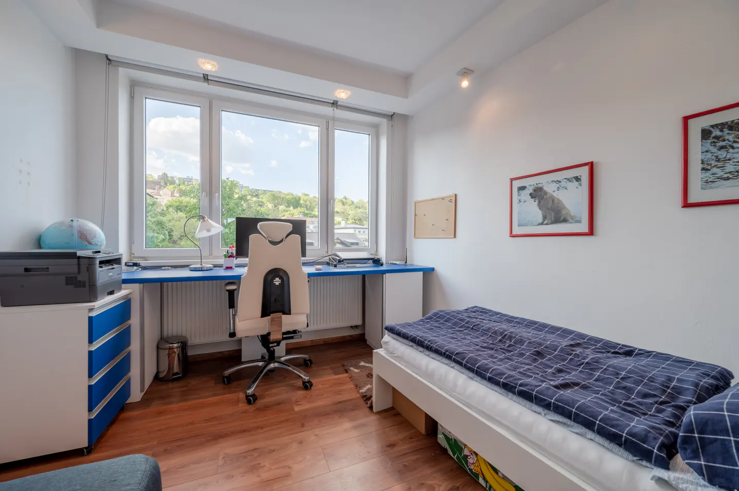 Bedroom with a desk, chair, and single bed. The desk is blue, and the chair is beige. There are framed pictures on the white wall. A large window overlooks greenery.