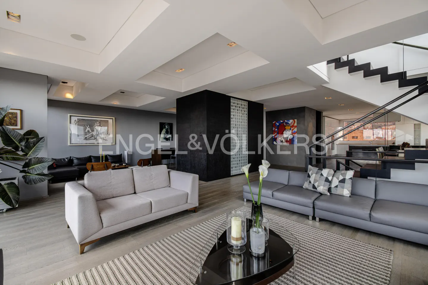 A modern living room with gray sofas, a black coffee table, and a staircase in the background. The walls are gray, and there is a large black column.