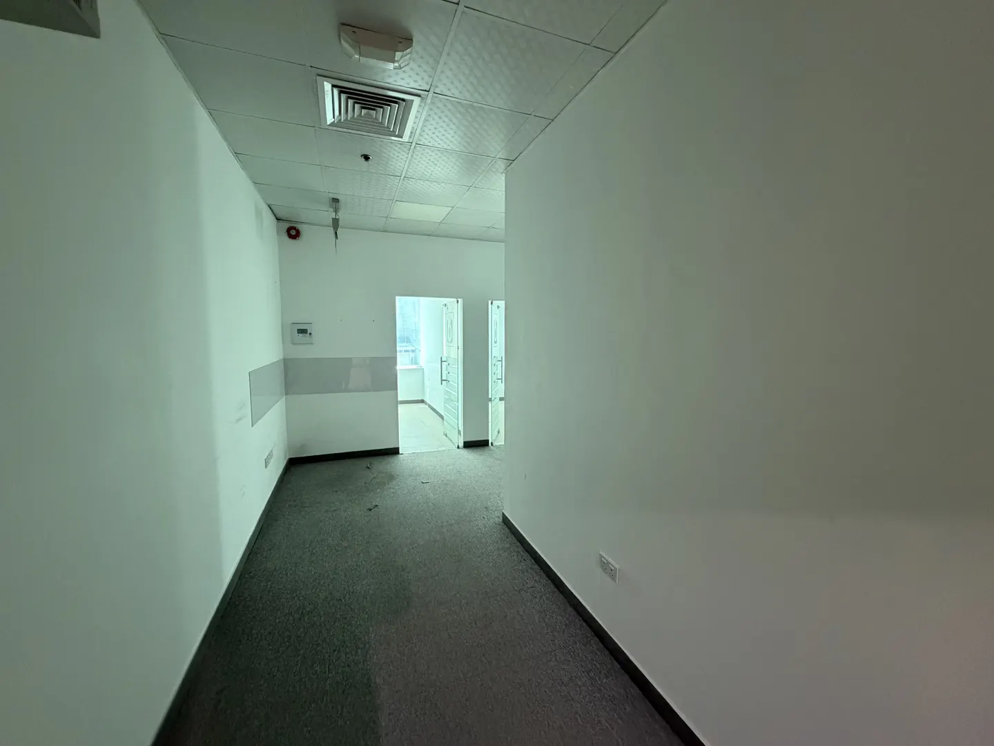 Empty office hallway with white walls, grey carpet, drop ceiling, and open doorways leading to a bright room.