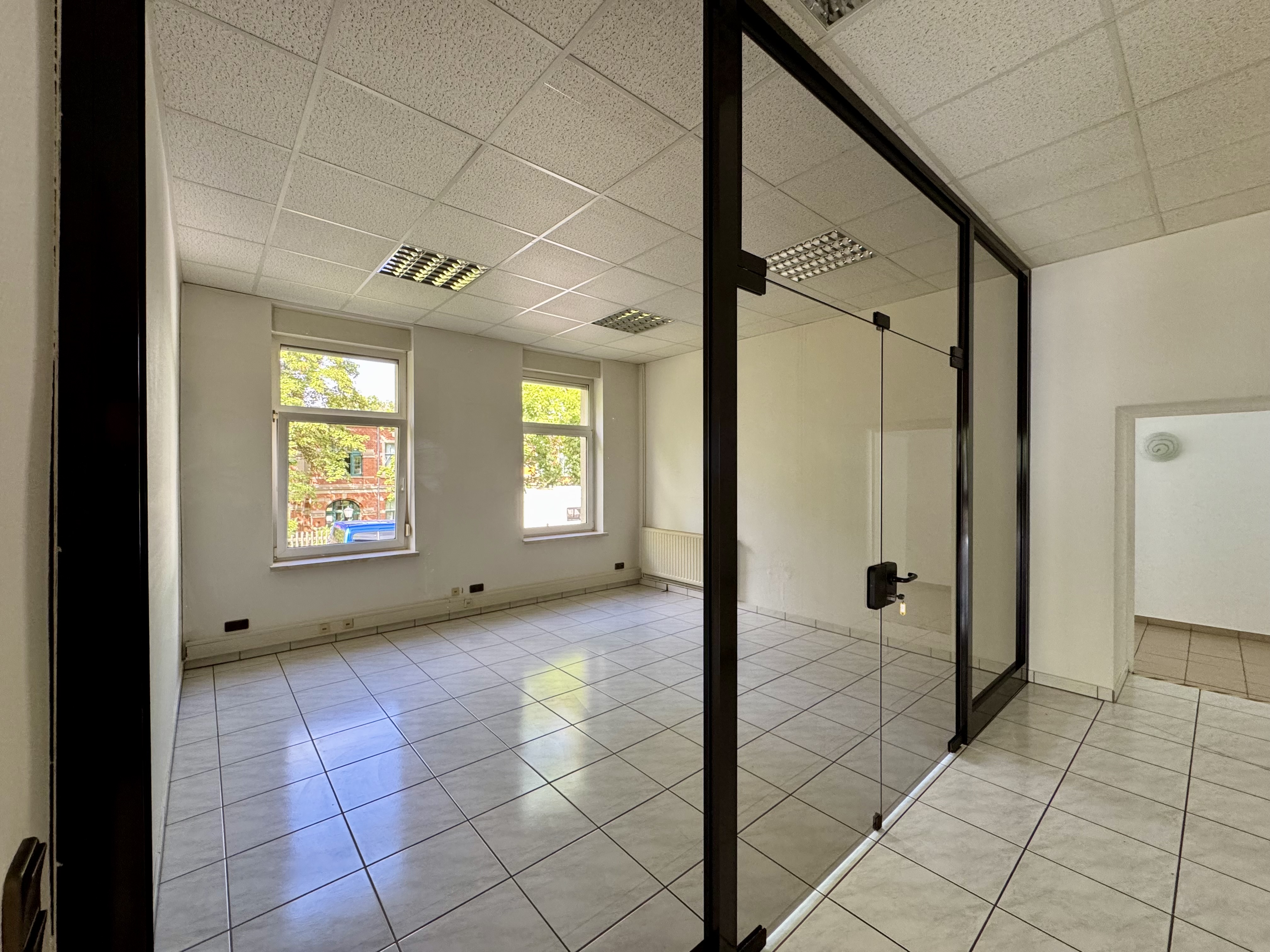 Empty office space with white walls, tile floors, and a glass-walled room divider. Windows overlook a building across the street.