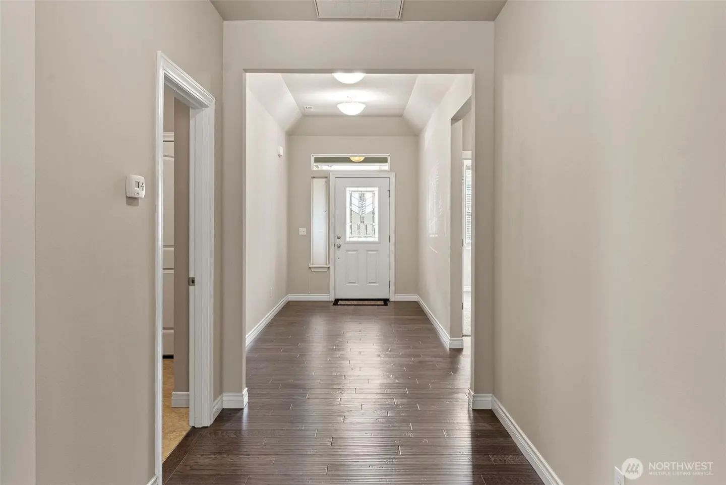 Entryway with dark wood floors, beige walls, and a white front door with glass panels. A doorway leads to another room on the left.