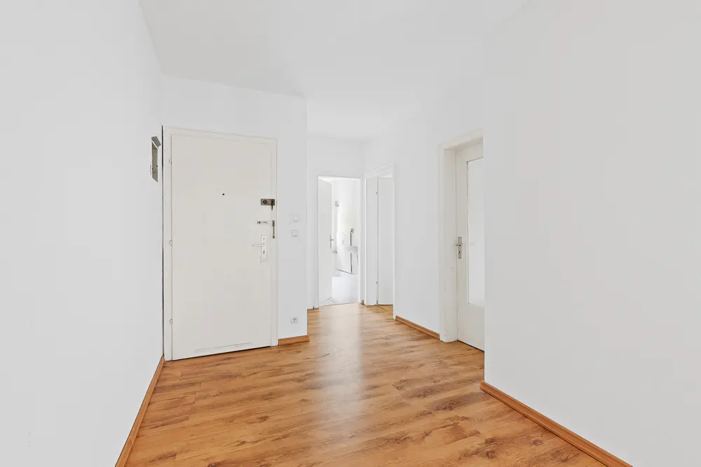 Bright hallway with wood floors and white walls. A white front door is on the left, and other doors lead to rooms.