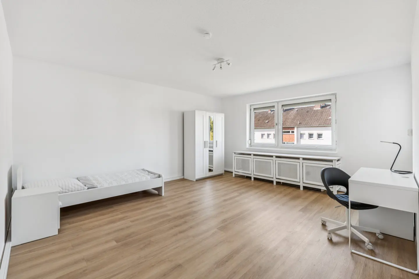 Bright, empty bedroom with a single bed, white wardrobe, desk, and chair on a wood floor. A window shows a view of a neighboring building.