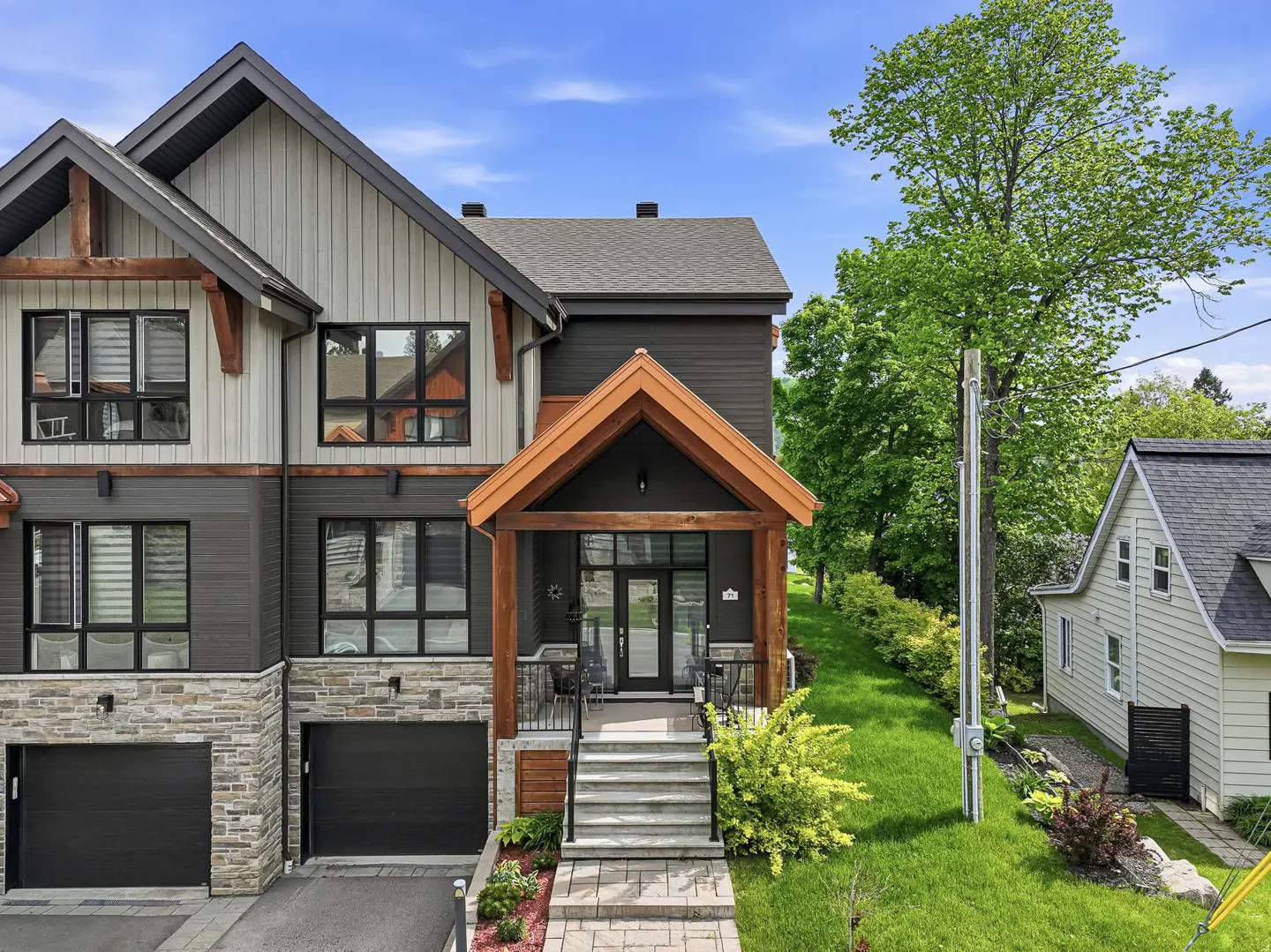 Modern two-story home with gray and beige siding, stone accents, a wooden porch, and two black garage doors.