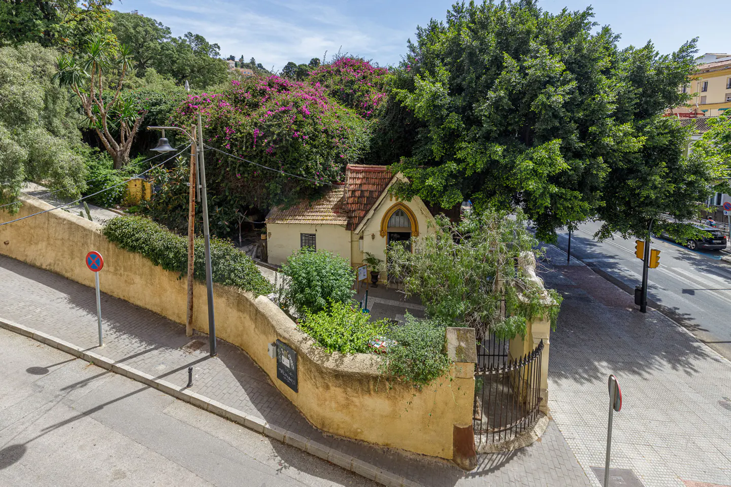 A small, cream-colored cottage with a tiled roof sits behind a curved yellow stone wall, surrounded by lush green trees.