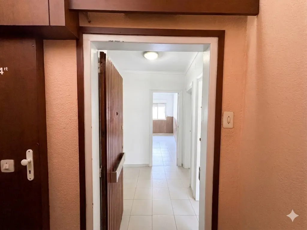 A view through a doorway into a bright hallway with white tile floors and white walls. A brown wooden door is partially open.