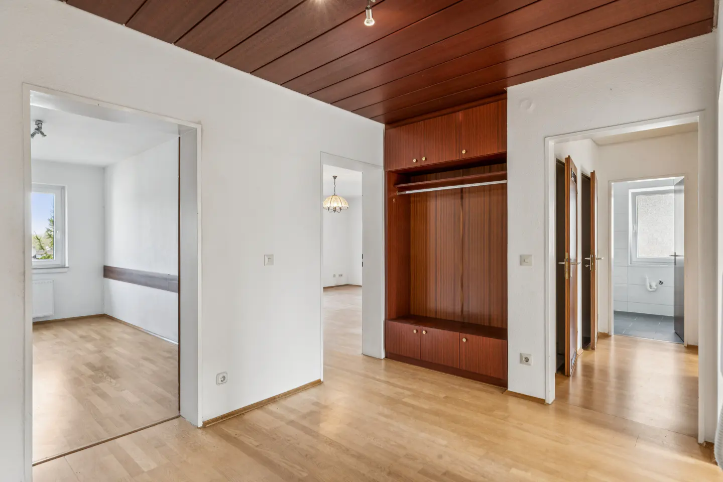 Interior view of a home with light wood floors, white walls, and a dark wood paneled ceiling. A built-in wood closet is on the right.