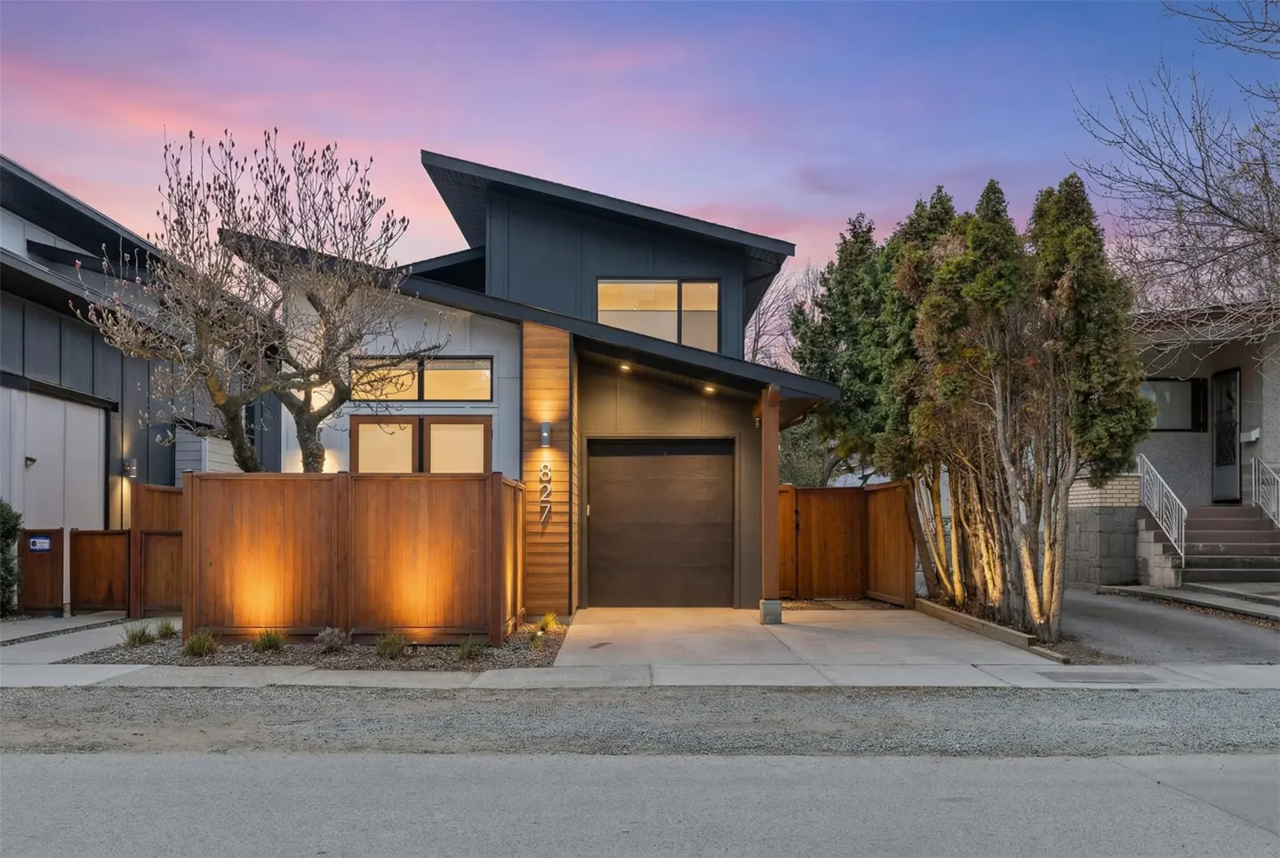 Modern home with a slanted roof, gray siding, and a brown garage door. A wooden fence surrounds the property. The sky is a mix of pink and purple.