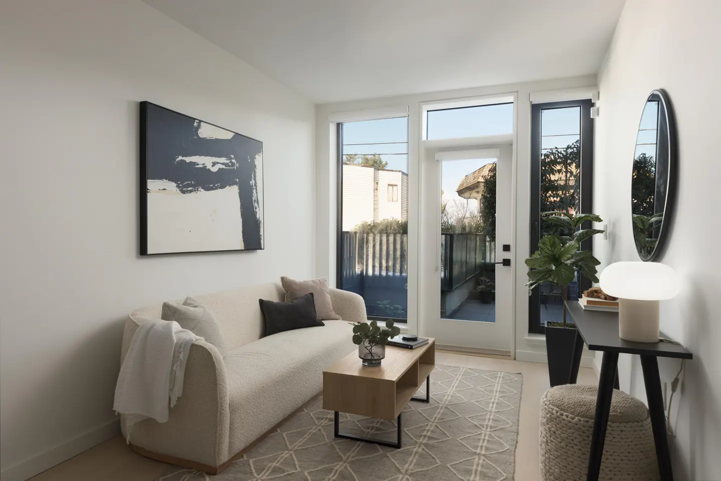 Bright living room with white walls, a cream sofa, and a geometric rug. A black and white abstract painting hangs above the sofa. A glass door leads to a patio.