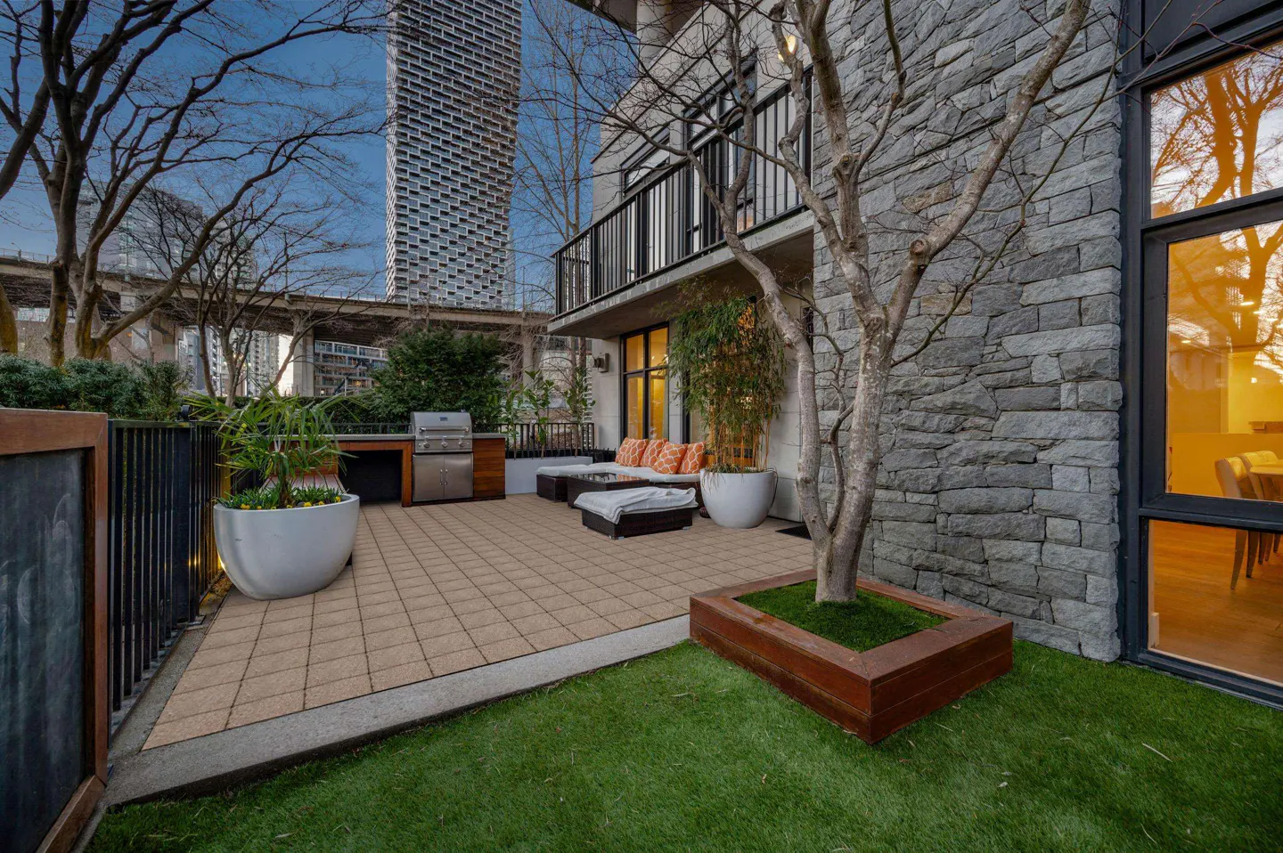 Outdoor patio with stone wall, grill, and seating area. A tree grows in a wooden planter box on the green lawn. A tall building is in the background.