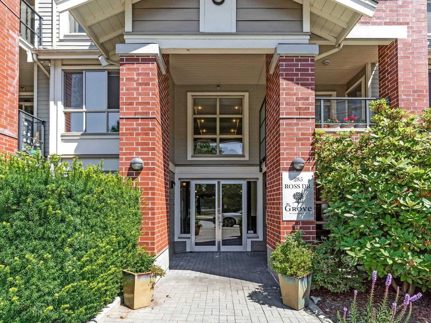 Exterior view of "The Grove" apartments at 285 Ross Dr. Brick pillars frame the glass doors and address sign. Green bushes flank the entrance.