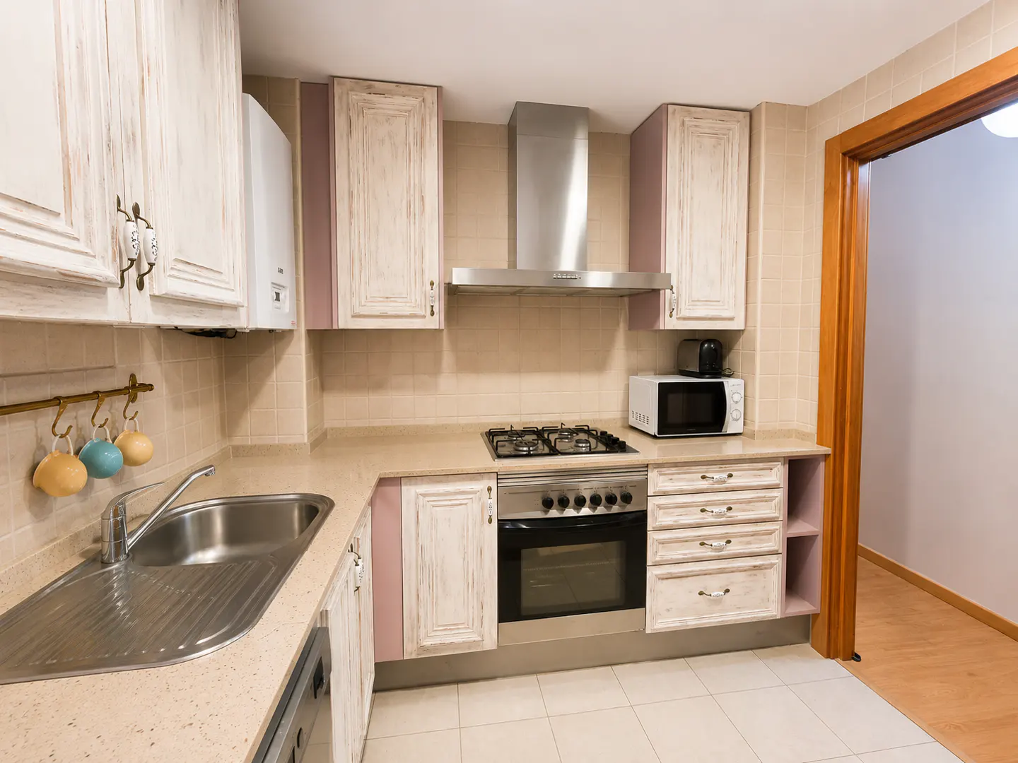 A well-lit kitchen with white and pink cabinets, stainless steel appliances, and a beige countertop. A doorway is visible on the right.
