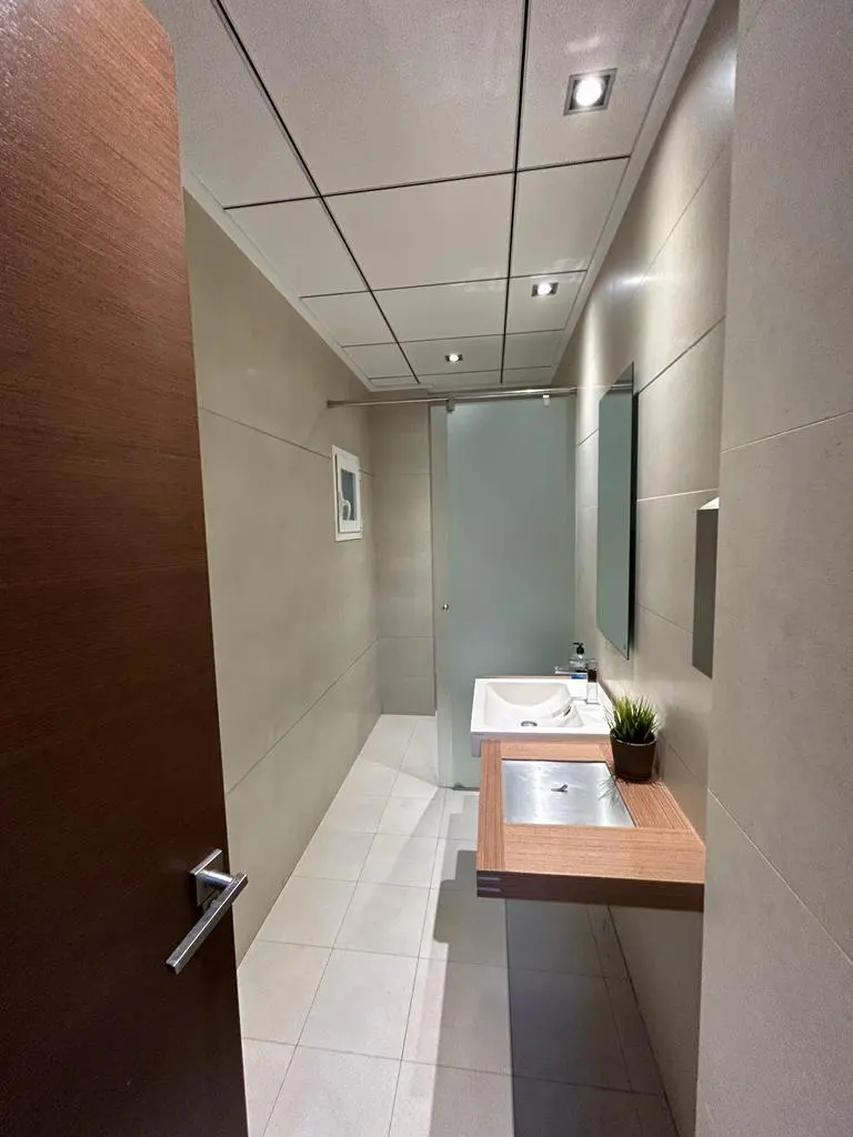Modern bathroom with a wood door, white tile floor, and a floating wood vanity with a white sink. A shower is visible in the background.