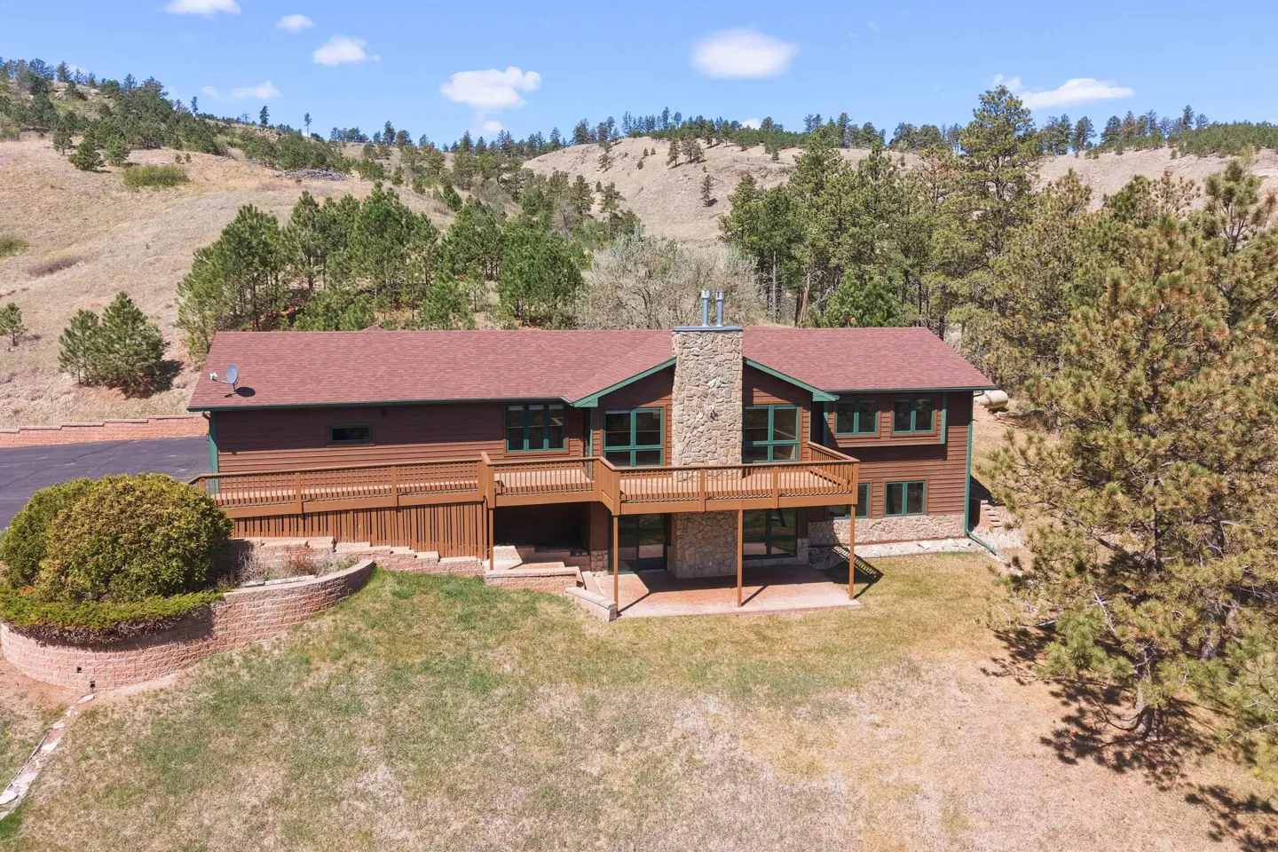 A brown, two-story house with a stone chimney, a deck, and a red roof sits on a grassy lawn with trees and a hill in the background.