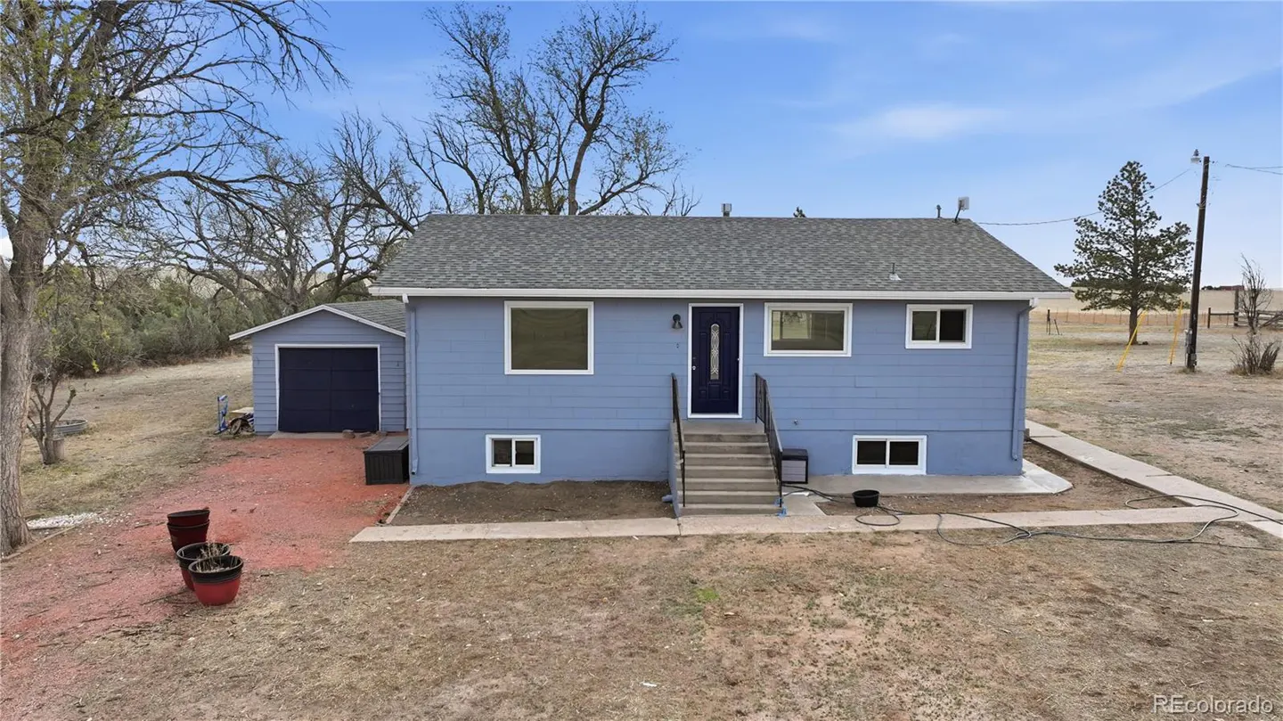 Blue single-story house with a dark front door, attached garage, and concrete steps in a rural, dry landscape.