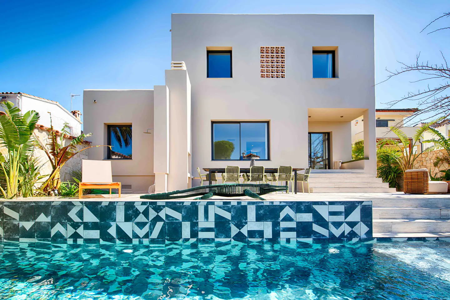 Modern white house with a pool. The pool has geometric tiles and turquoise water. A table and chairs sit on the patio.