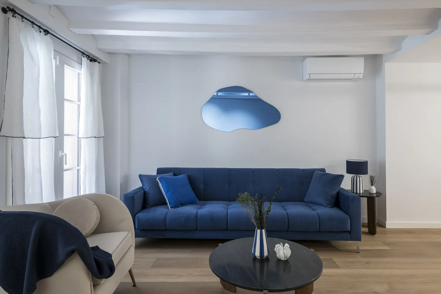 Bright living room with a blue velvet sofa, round coffee table, and a blue abstract mirror on a white wall.