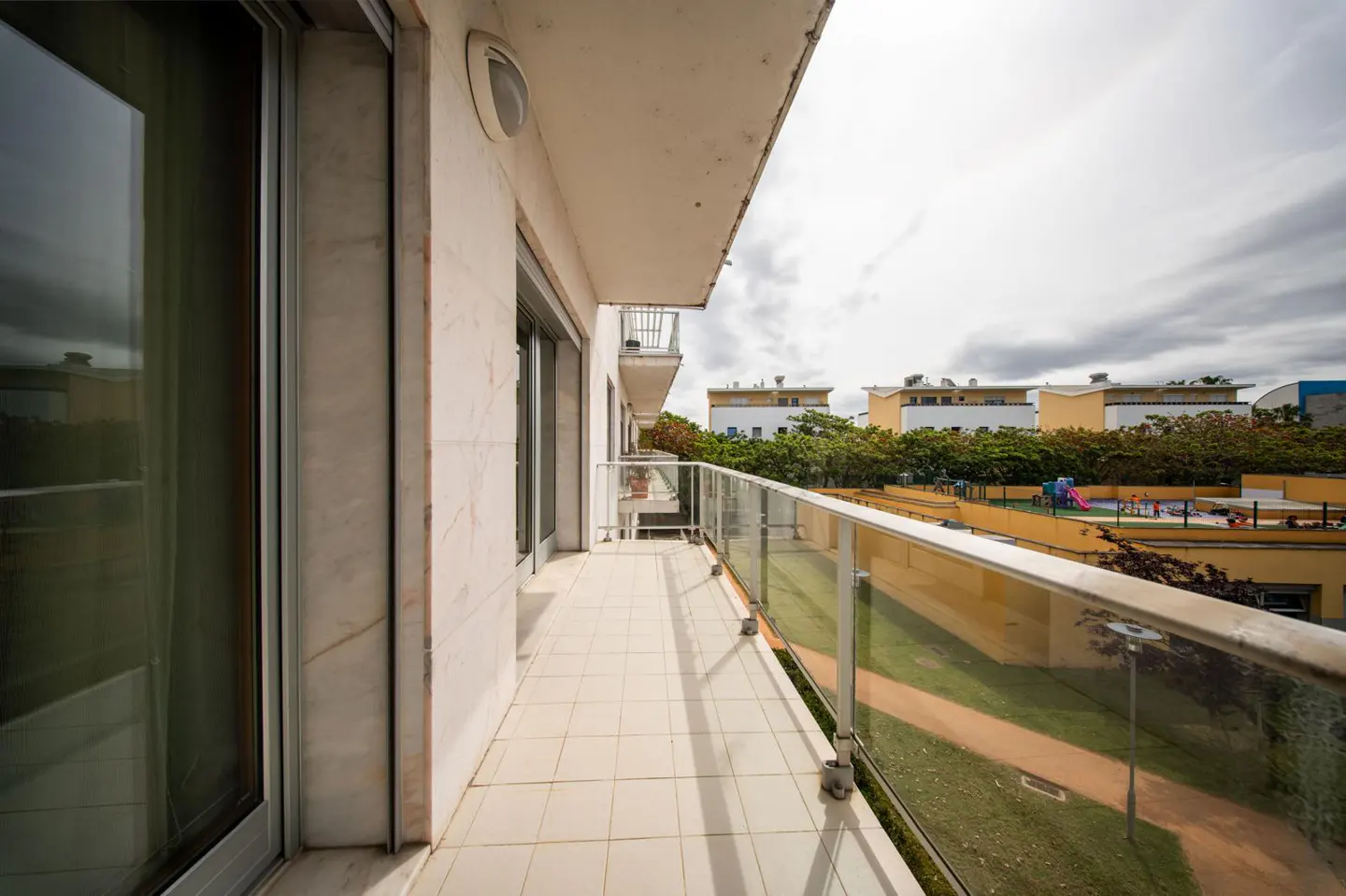 A long balcony with glass railings overlooks a playground and buildings under a cloudy sky.