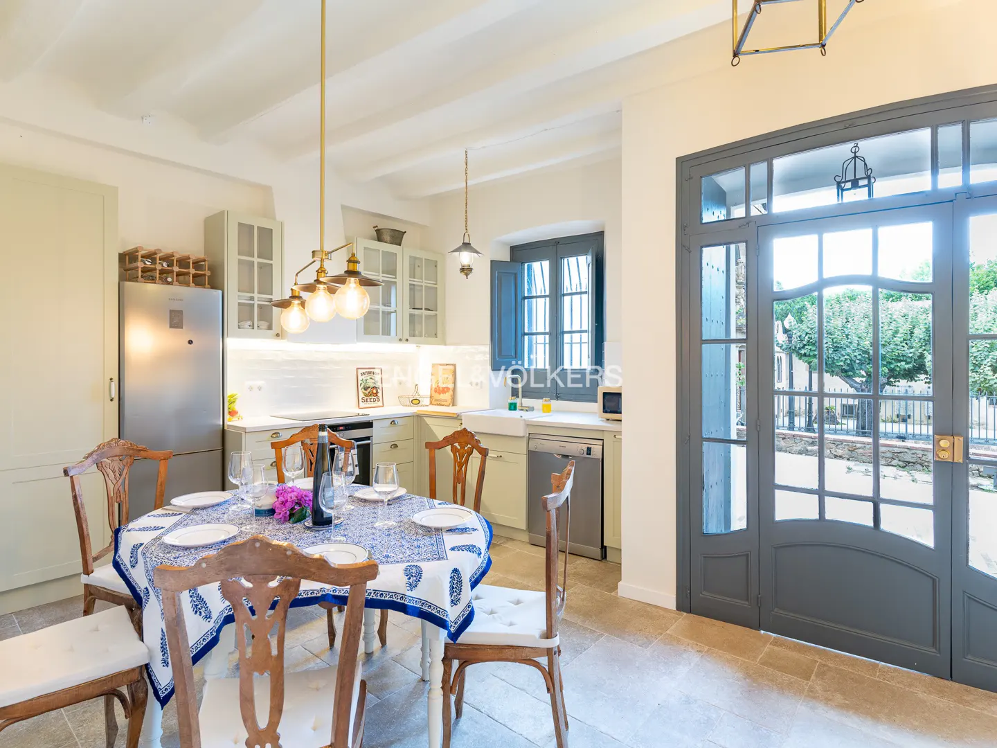 Bright kitchen with a round dining table, blue and white patterned tablecloth, wooden chairs, and a large glass-paned door.
