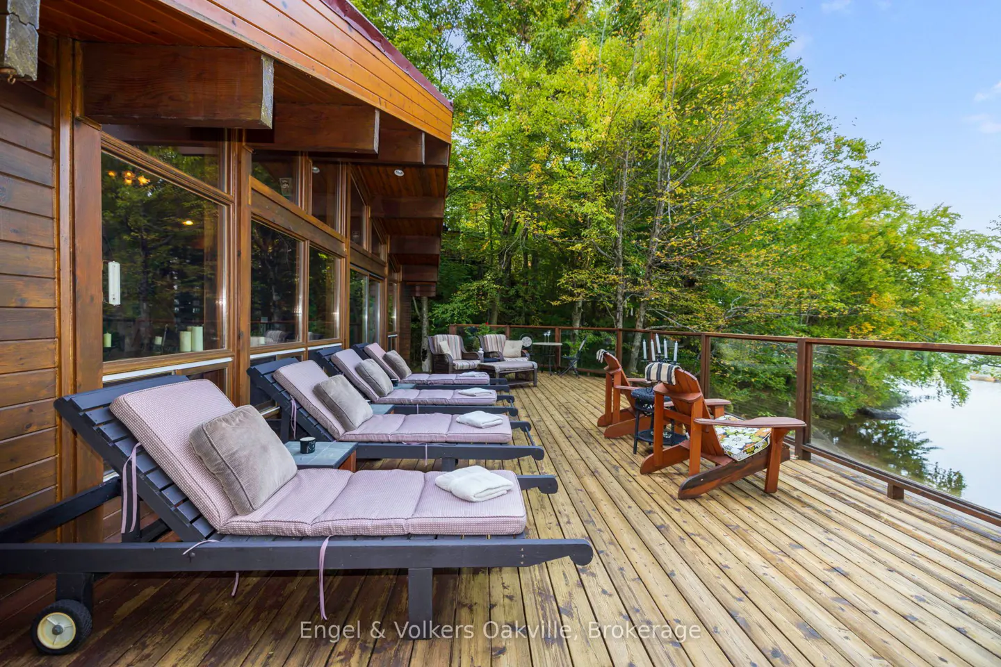 Wooden deck with lounge chairs and Adirondack chairs overlooking a lake surrounded by green trees.