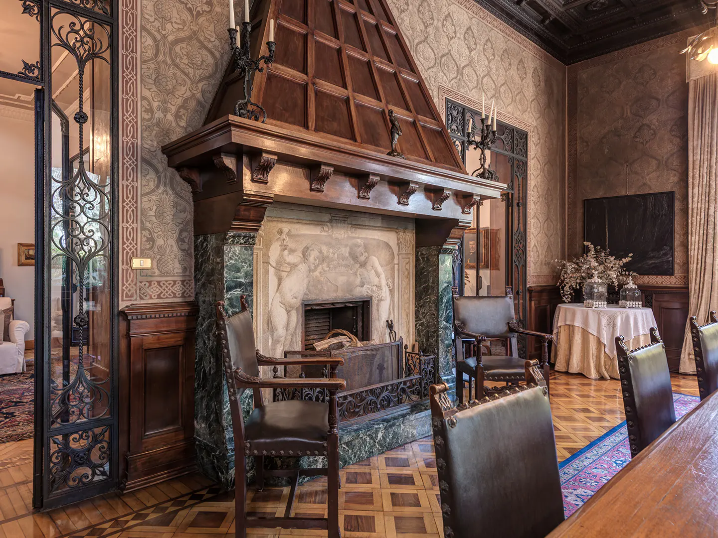 Ornate room with a large, dark wood fireplace featuring marble accents and a carved relief. Chairs are arranged around the fireplace.