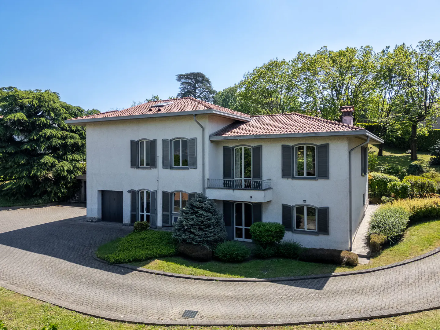 Two-story white house with gray shutters and a red tile roof, surrounded by green trees and a circular driveway.