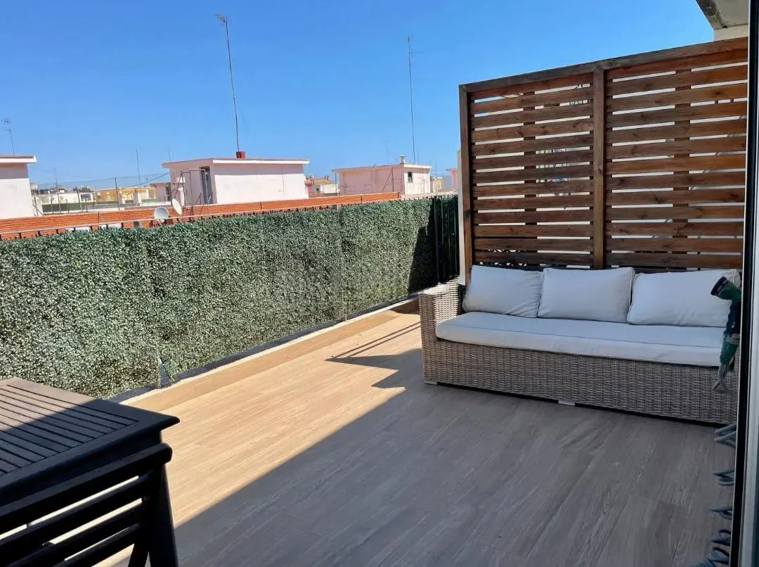 Outdoor patio with wicker sofa, wood screen, and green hedge. Blue sky in background.