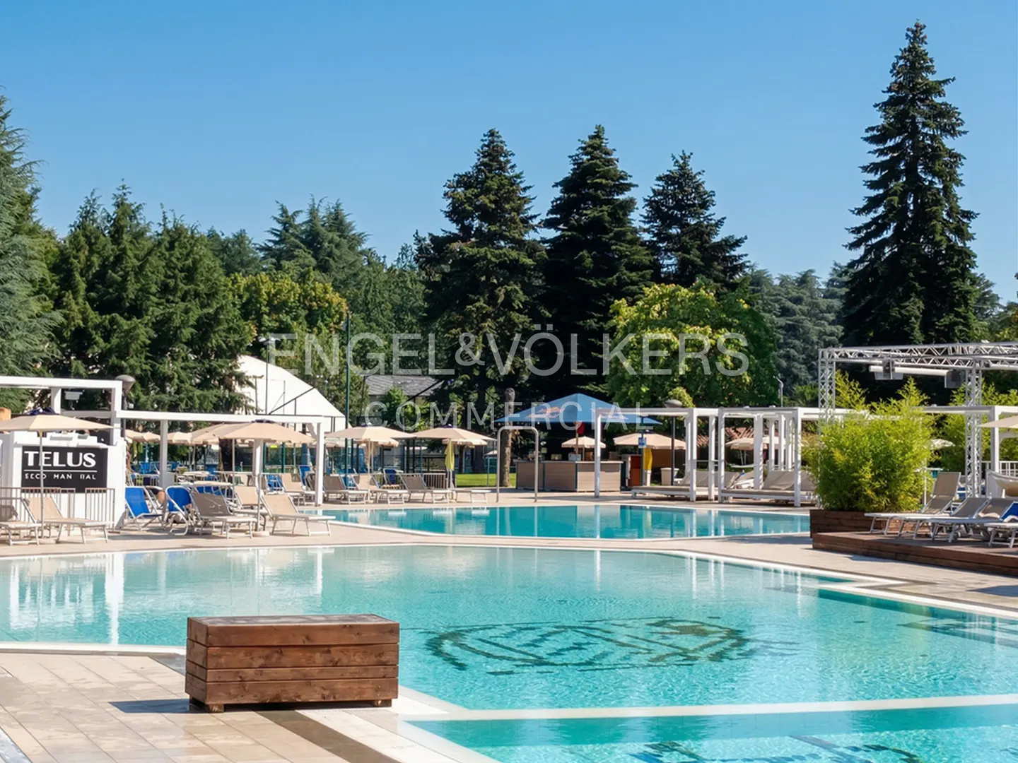 Outdoor pool area with lounge chairs, umbrellas, and cabanas. Trees and blue sky in the background.