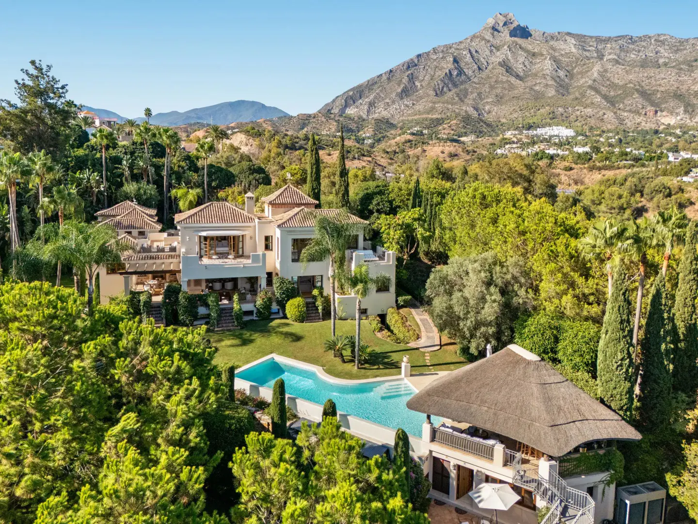 Aerial view of a large, light-colored villa with a pool, surrounded by lush greenery and mountains in the background.