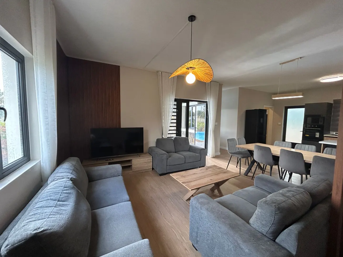 Modern open-plan living room with grey sofas, wooden coffee table, dining area, and a view of a pool through glass doors.