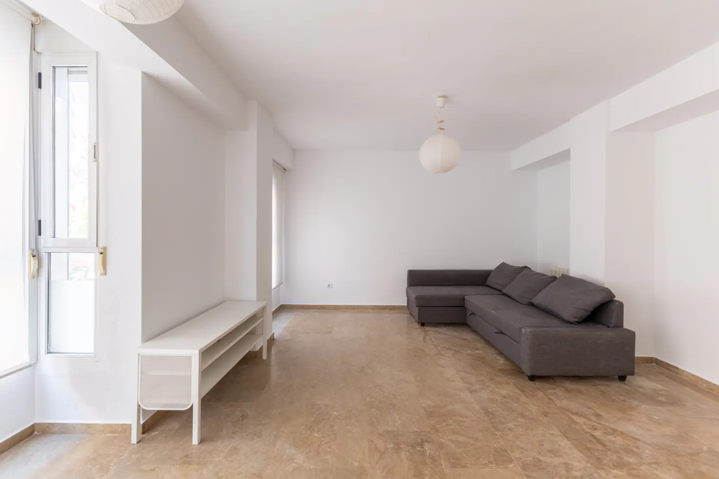 Bright, empty living room with marble floors, white walls, and a gray sectional sofa. A white console table sits near a window.