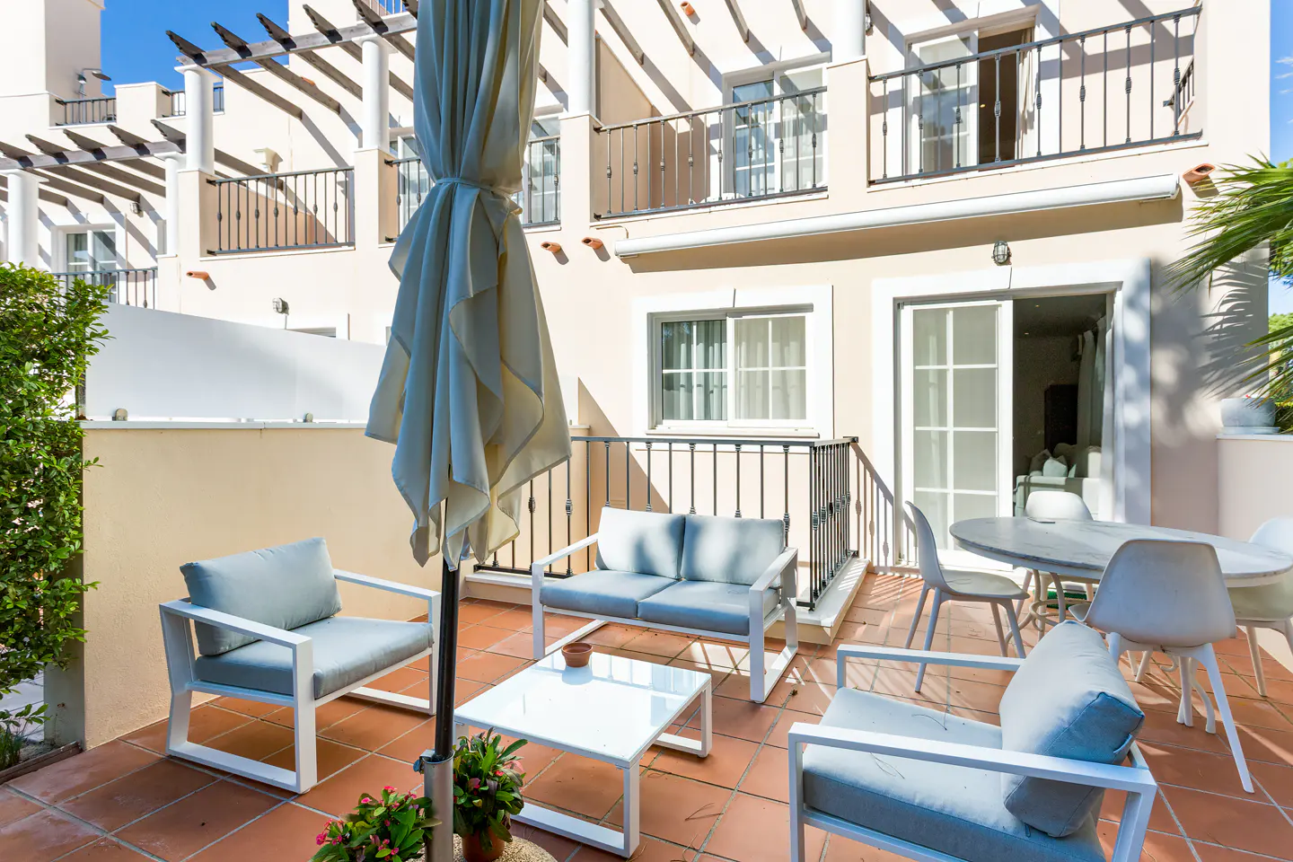 Outdoor patio with white furniture, including chairs, a loveseat, and a table. A closed umbrella stands nearby. The patio has terracotta tiles. Cream-colored building in the background.