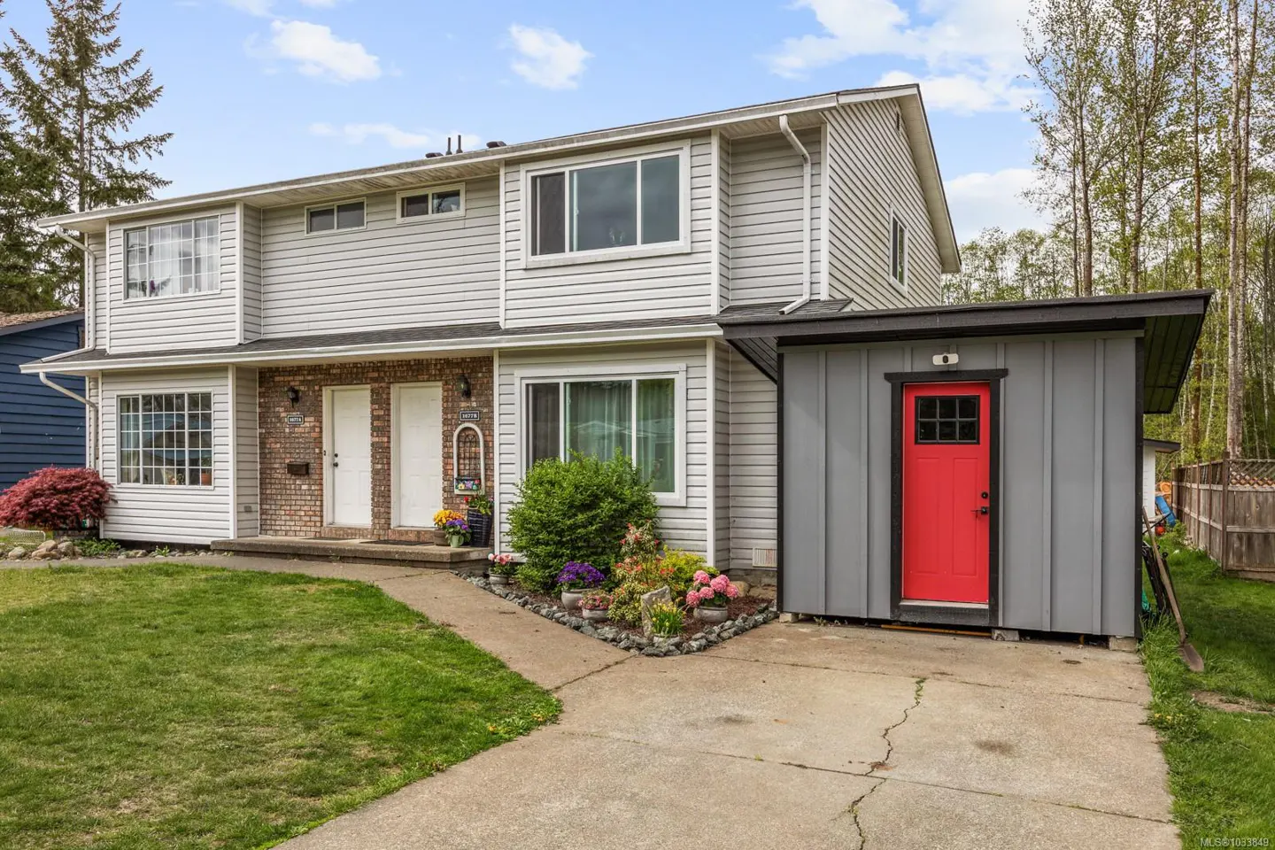 Two-story duplex with light gray siding, brick accents, white doors, and a gray shed with a red door. Lawn and flower beds in front.