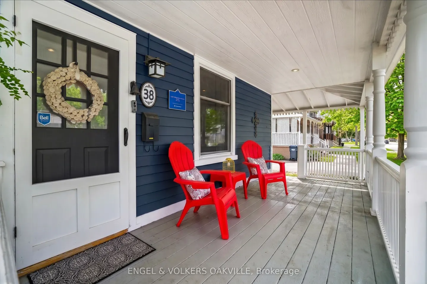Covered porch with two red Adirondack chairs, a small red table, and a white door with a wreath on a blue house.