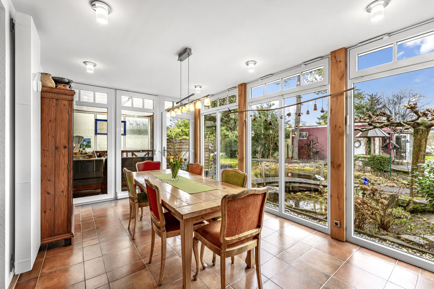 Bright dining room with a wooden table, colorful chairs, and a view of a lush garden through large windows.