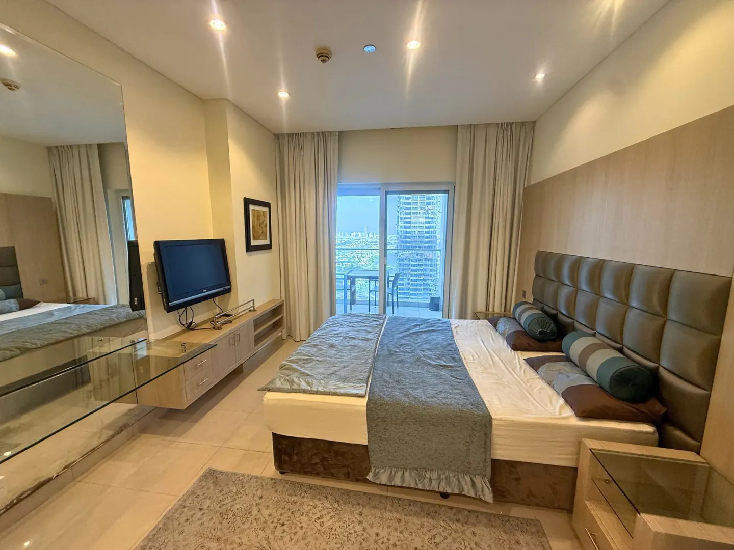 Bedroom with a large bed, blue blanket, and balcony with city views. A TV sits on a light wood cabinet. A large mirror reflects the room.