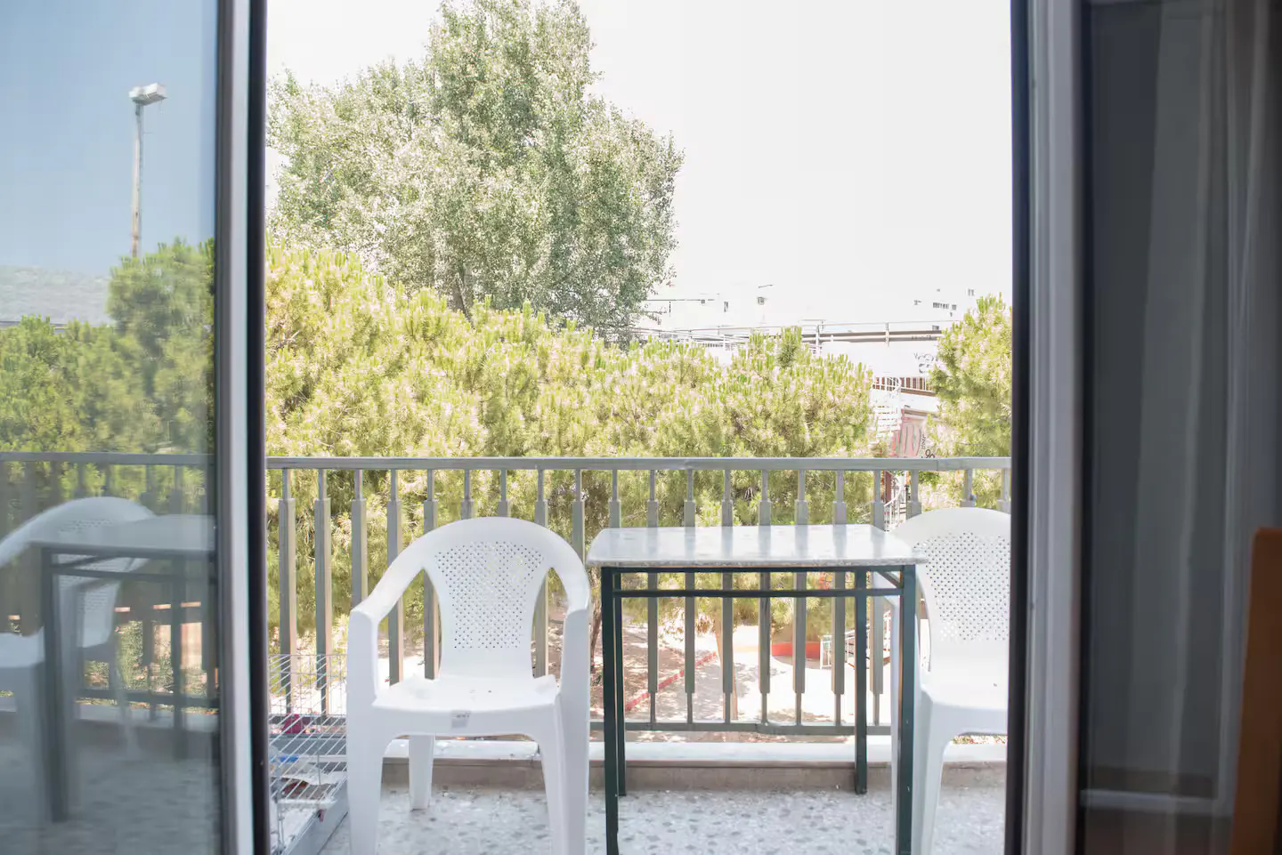 Balcony view with white plastic chairs and a small table, overlooking green trees and a cityscape. Sliding glass doors frame the scene.