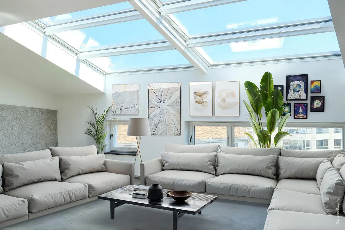 Bright living room with skylights, a large gray sectional sofa, and a marble coffee table. Artwork and plants decorate the space.