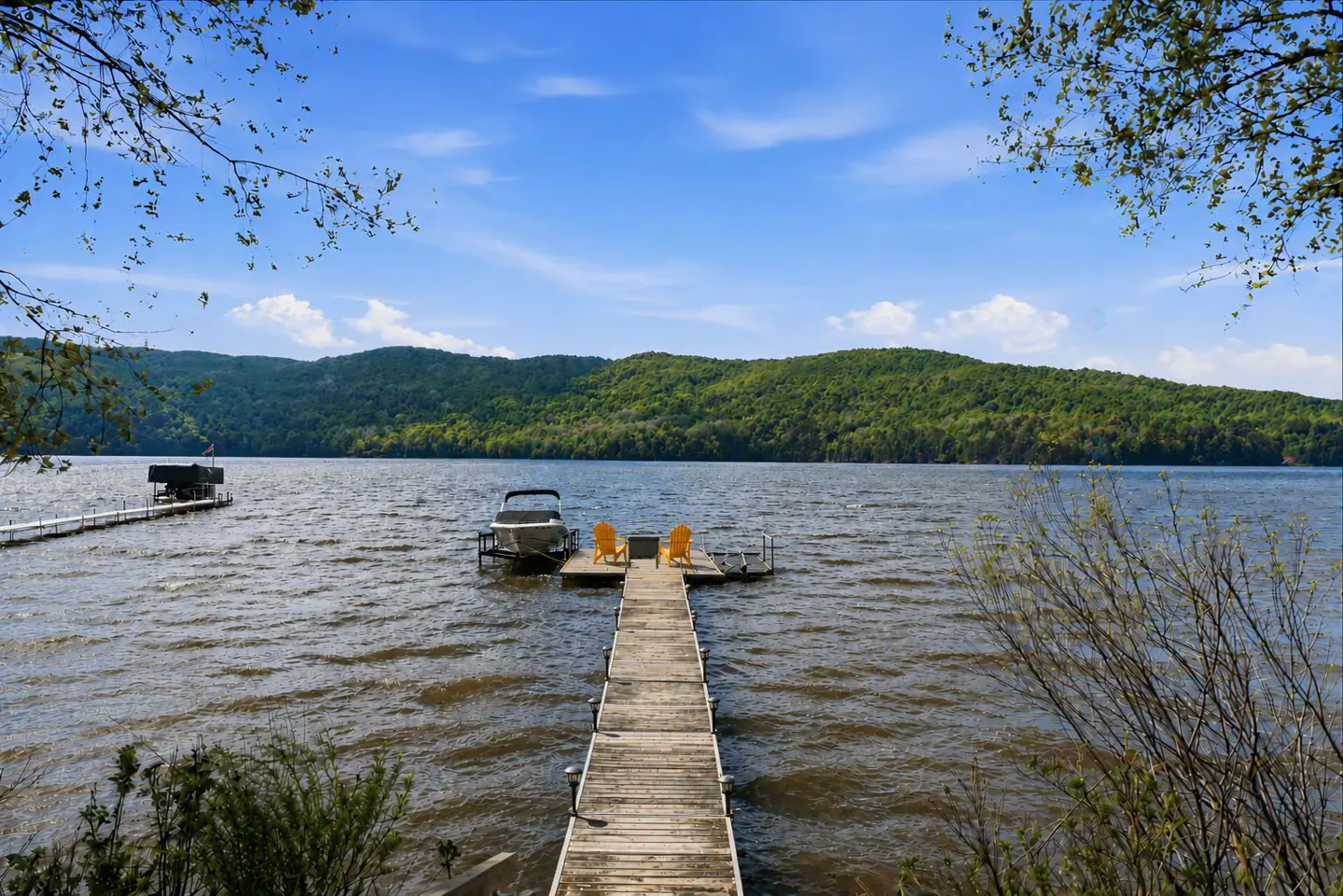 Lake view with a wooden dock, two yellow chairs, and a boat. Green hills and a blue sky with clouds in the background.