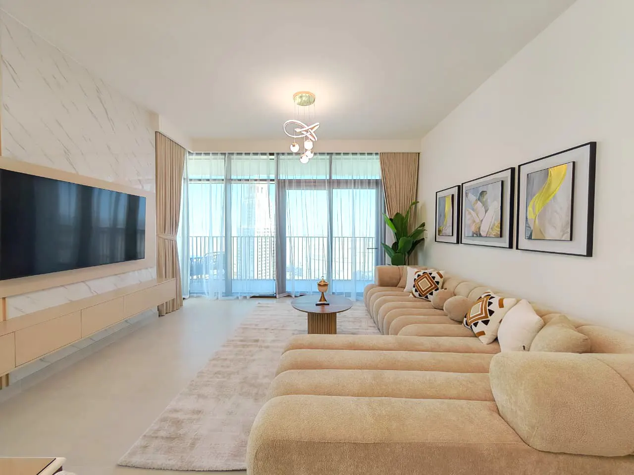 Bright living room with a beige sectional sofa, round coffee table, and large windows with sheer curtains. Artwork hangs on the white wall.