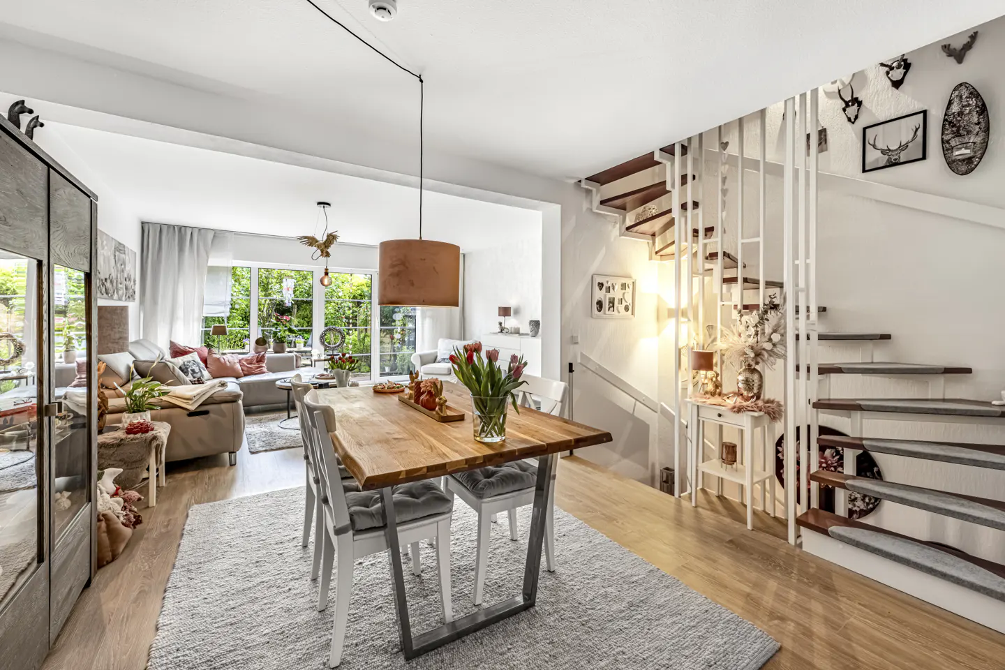Bright, open-concept living and dining area with wood table, white chairs, and a gray rug. Stairs with white railings lead to the upper level. Sofa visible in the background.
